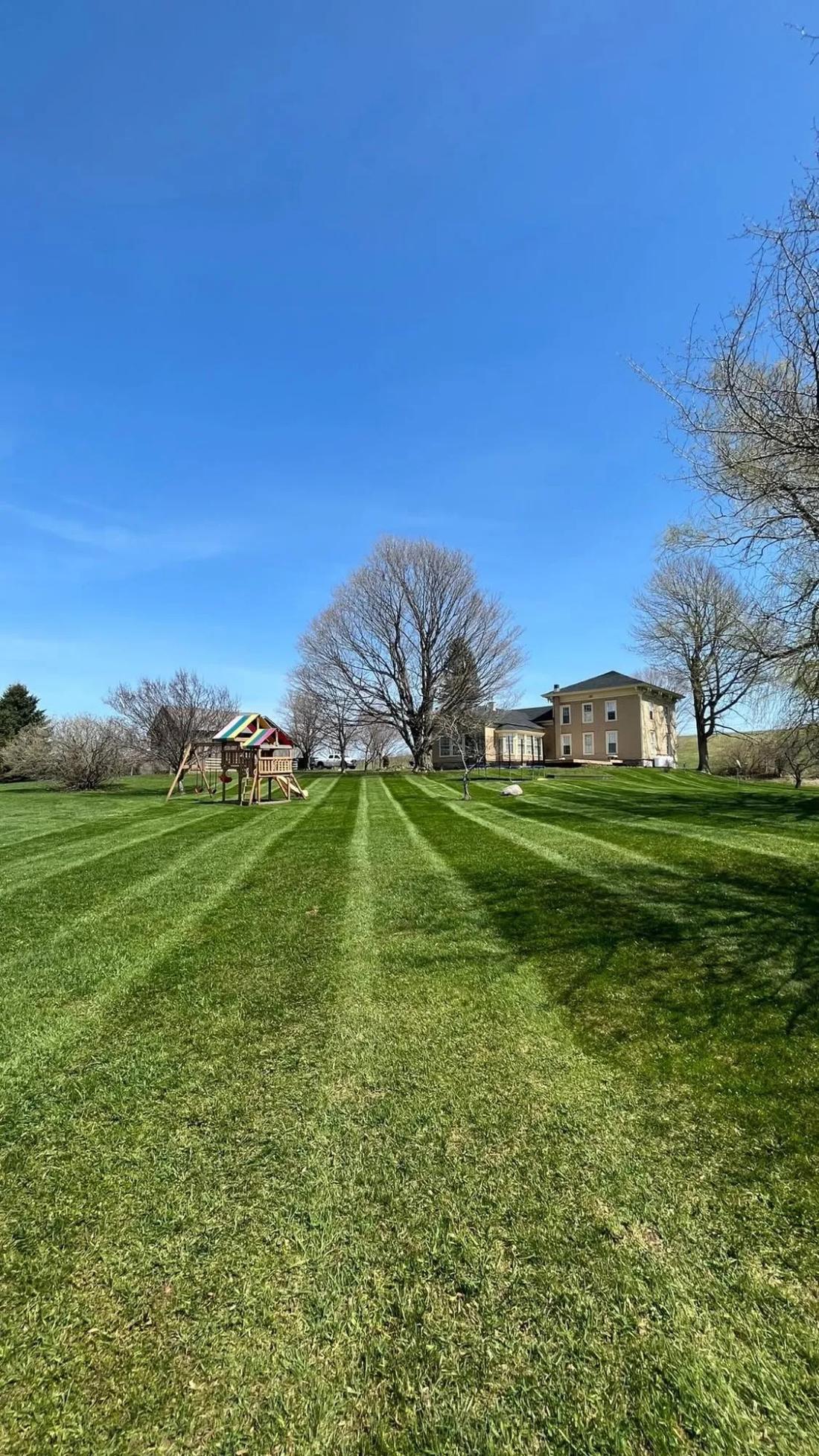 A large lawn mowed in clean straight stripes running toward a farmhouse, with a play set off to one side